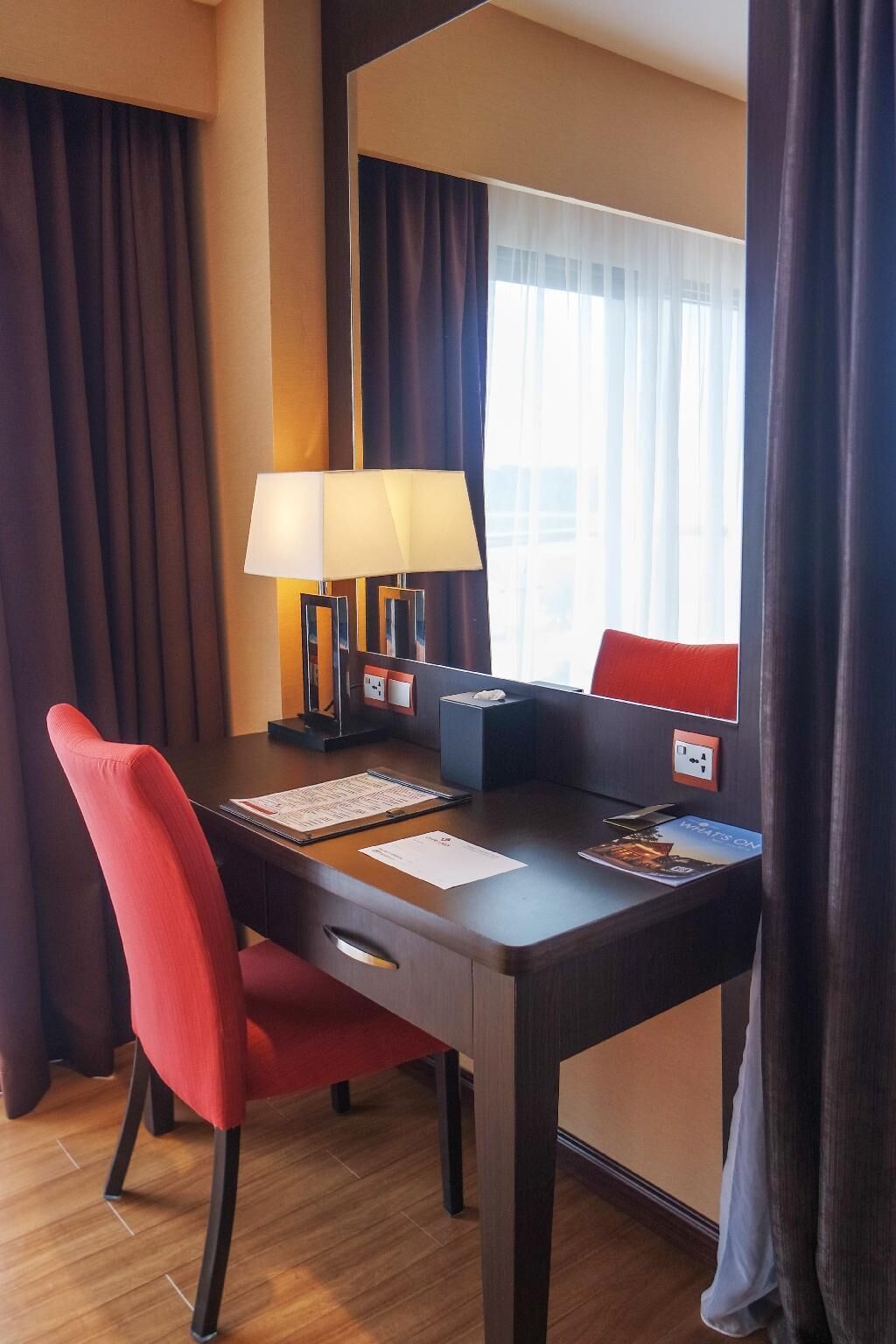 Une chambre d’hôtel avec un bureau en bois, une chaise rouge, une lampe, une fenêtre avec des rideaux et une vue sur la ville.