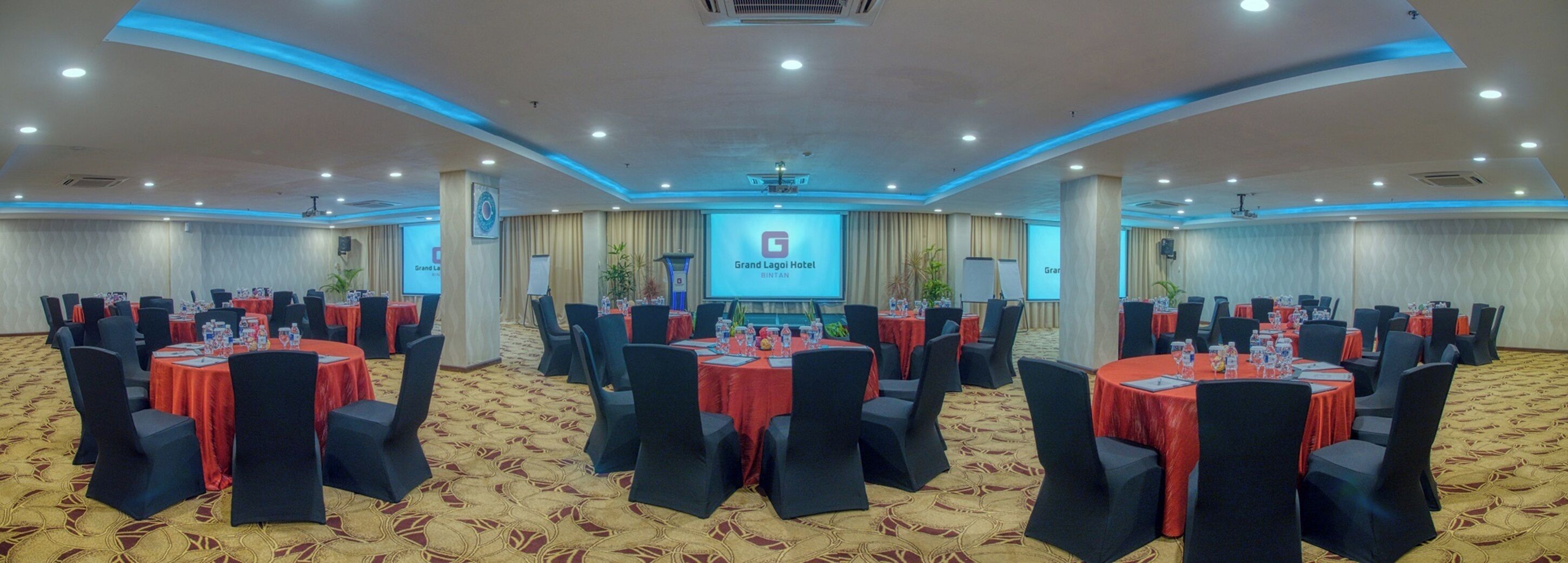 A conference room with multiple tables and chairs, a large screen at the front, and a carpeted floor.