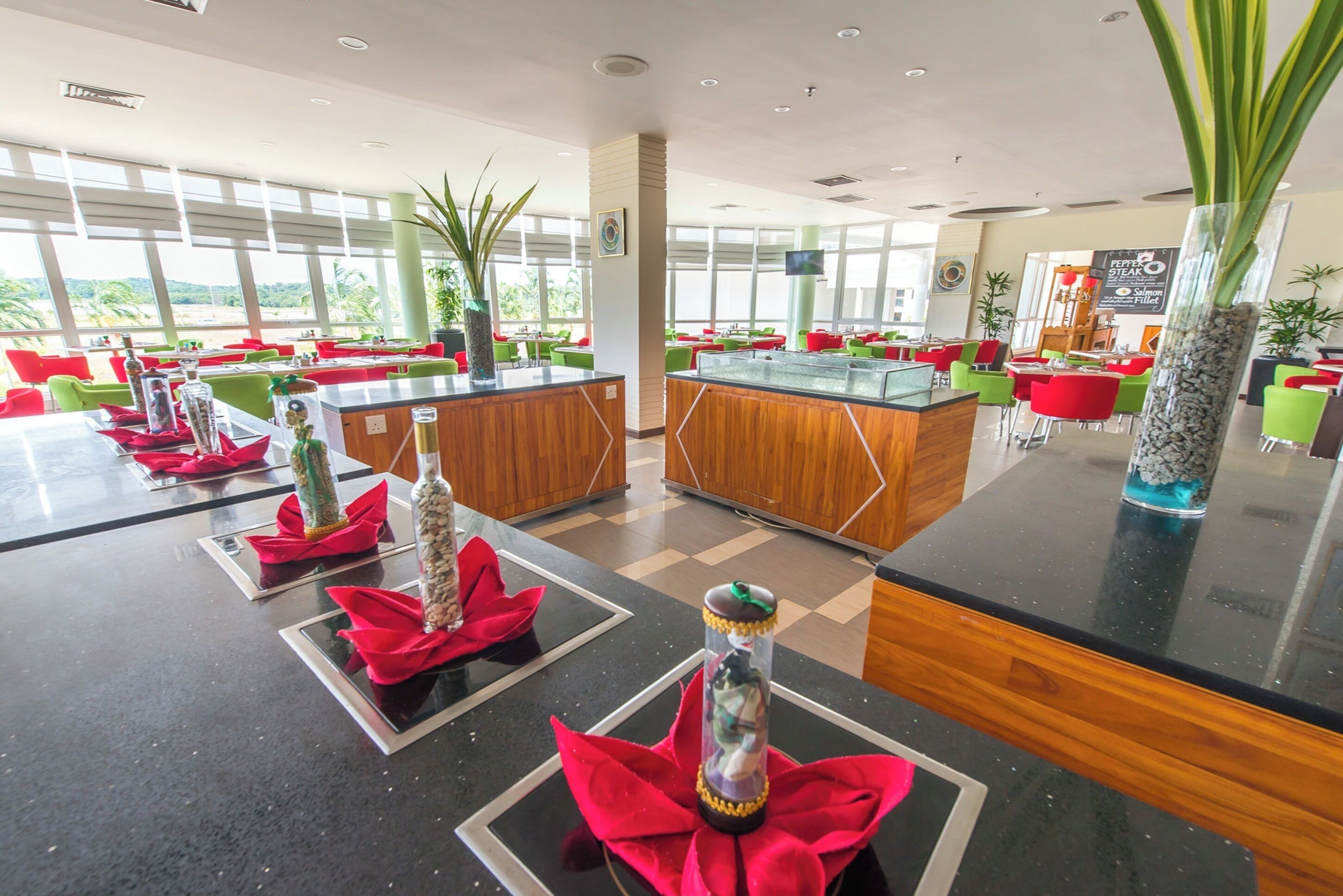 Um hall de hotel moderno com uma recepção central, áreas de estar com cadeiras vermelhas e verdes, e grandes janelas com vista para o exterior.