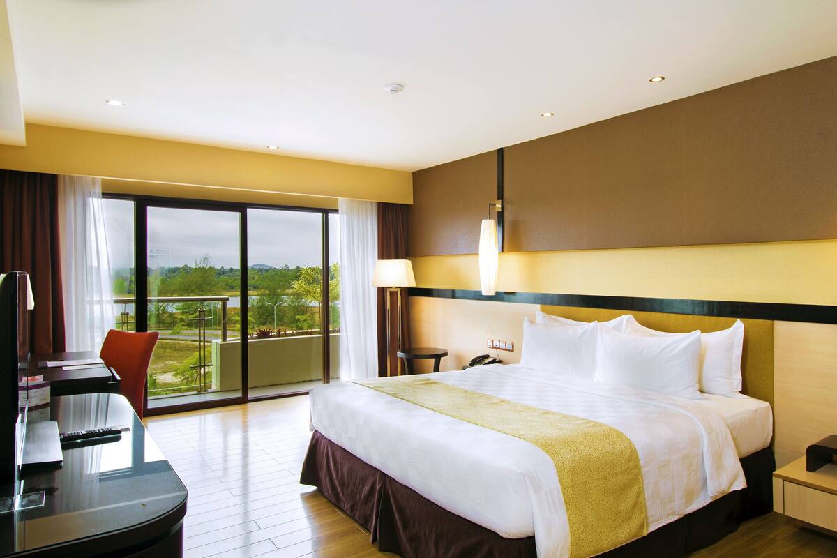A hotel room with a large bed, a desk, and a chair. There is a balcony with a view of greenery.