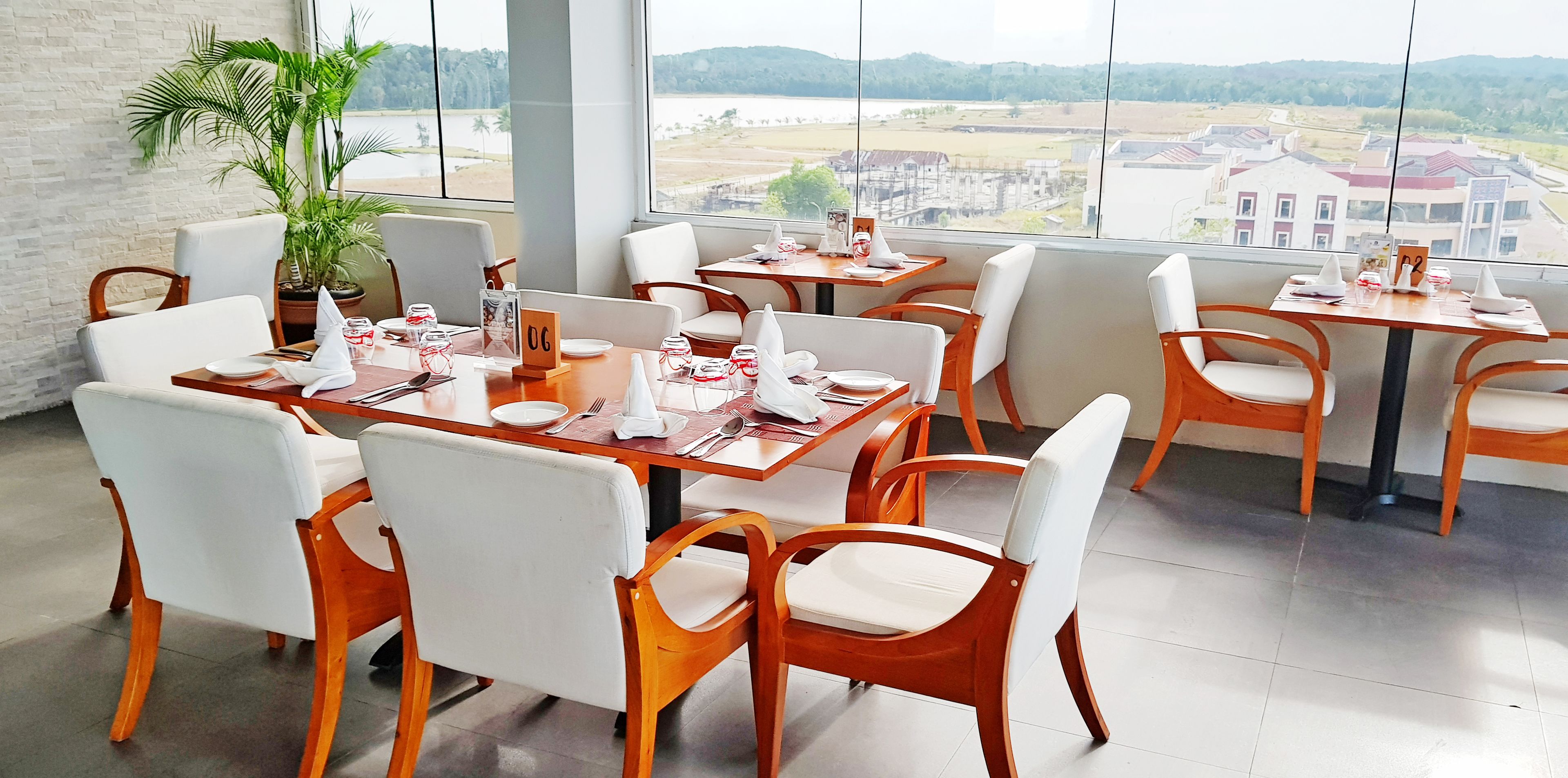 Un restaurante con mesas de madera y sillas blancas, grandes ventanales con vistas a edificios y zonas verdes.