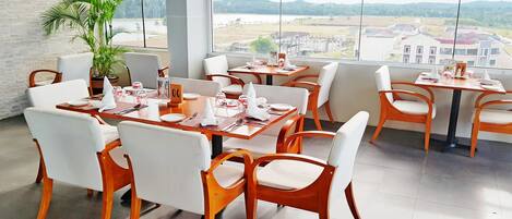Un restaurante con mesas de madera y sillas blancas, grandes ventanales con vistas a edificios y zonas verdes.