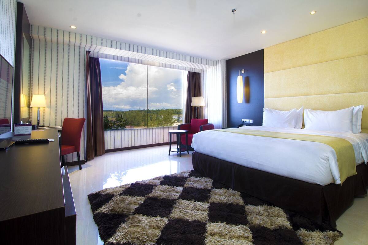 A hotel room with a large bed, a desk with a computer, a chair, and a view of the outdoors through a large window.