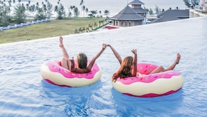 Two people floating on pink and white inflatable tubes in a pool with a view of the ocean and palm trees in the background.