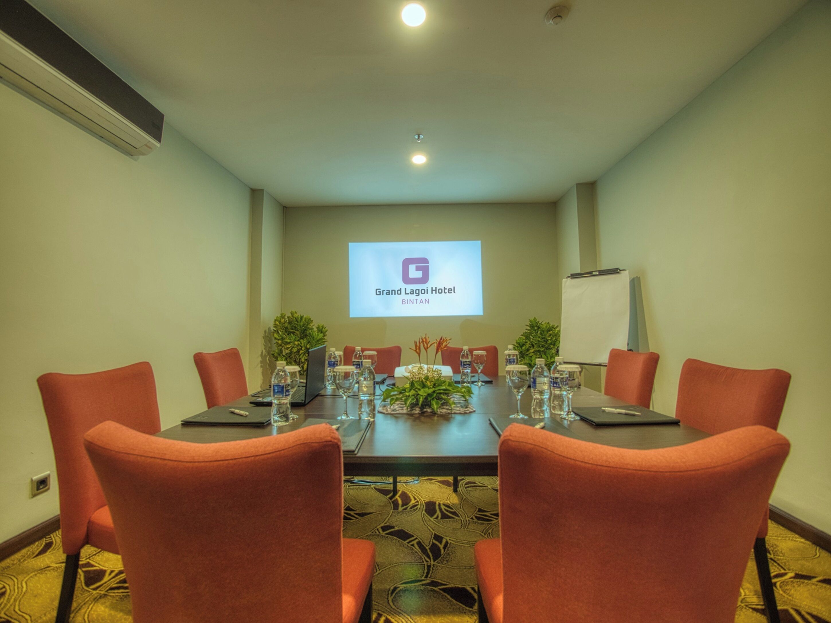 A conference room with a long table, six orange chairs, water bottles, and a laptop. A projector screen displays the Grand Lagoon Hotel logo.