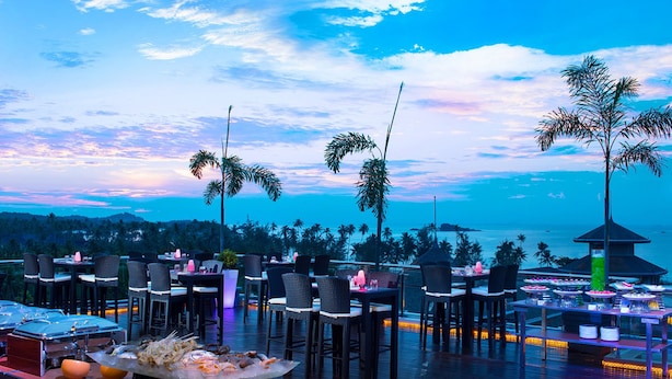An outdoor dining area with tables and chairs, overlooking a scenic ocean view with palm trees and a sunset.