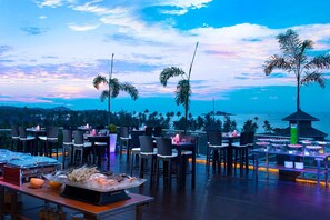 An outdoor dining area with tables and chairs, overlooking a scenic ocean view with palm trees and a sunset. - Grand Lagoi Hotel Bintan (Bintan)