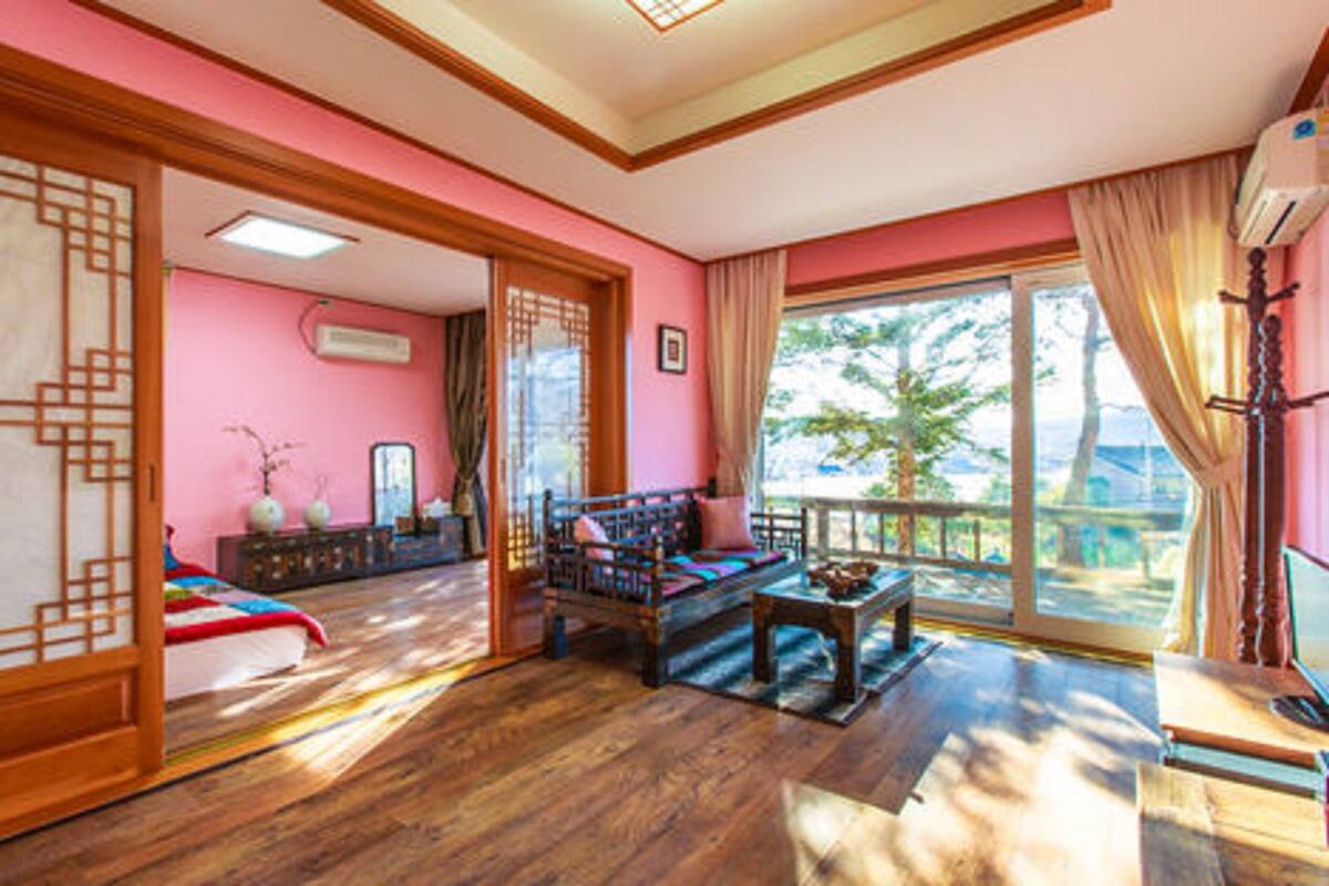 A room with a traditional design, featuring a wooden floor, a sofa, a table, and a large window with a view of trees.