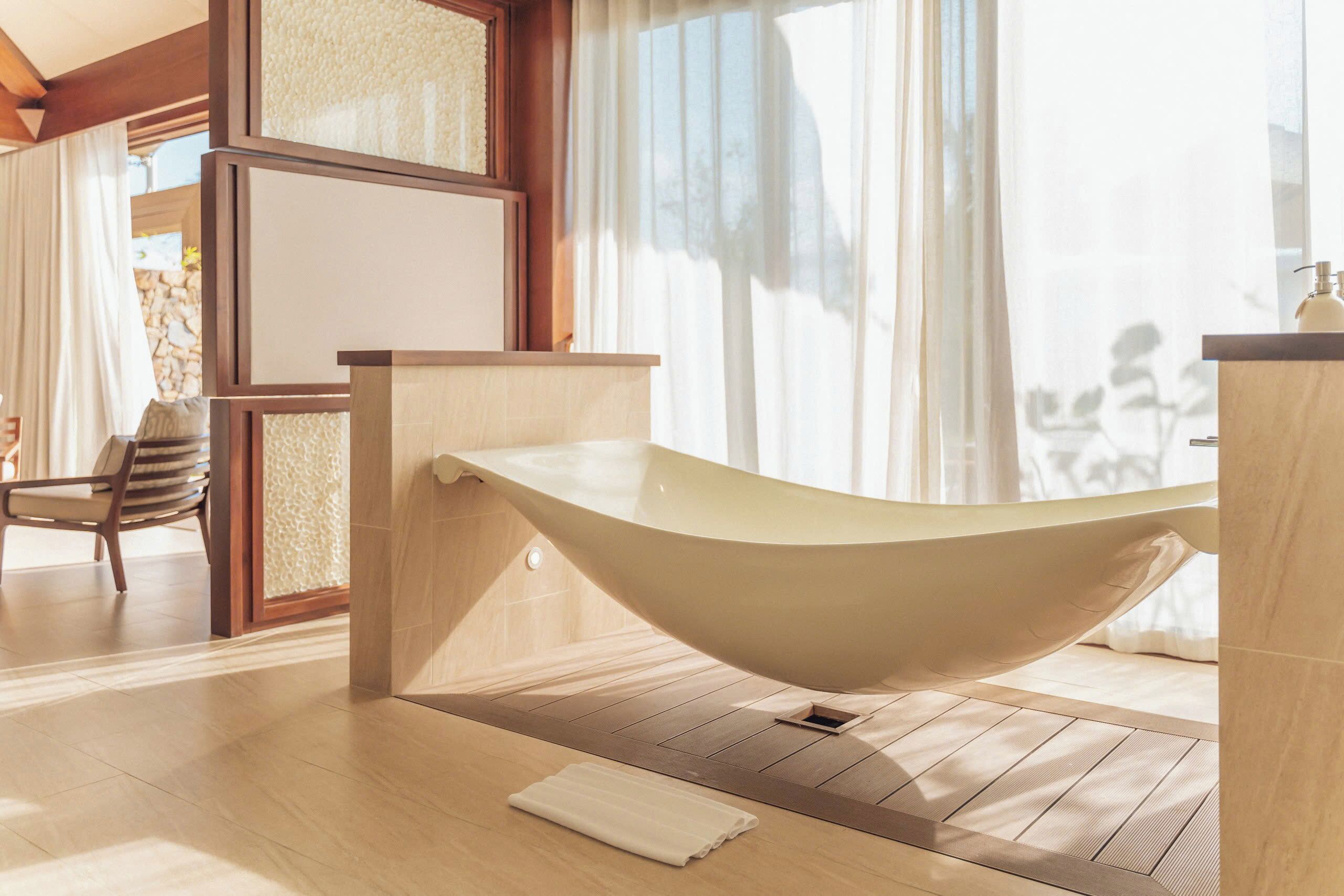 A modern bathroom with a freestanding bathtub, wooden flooring, and a large window with sheer curtains.