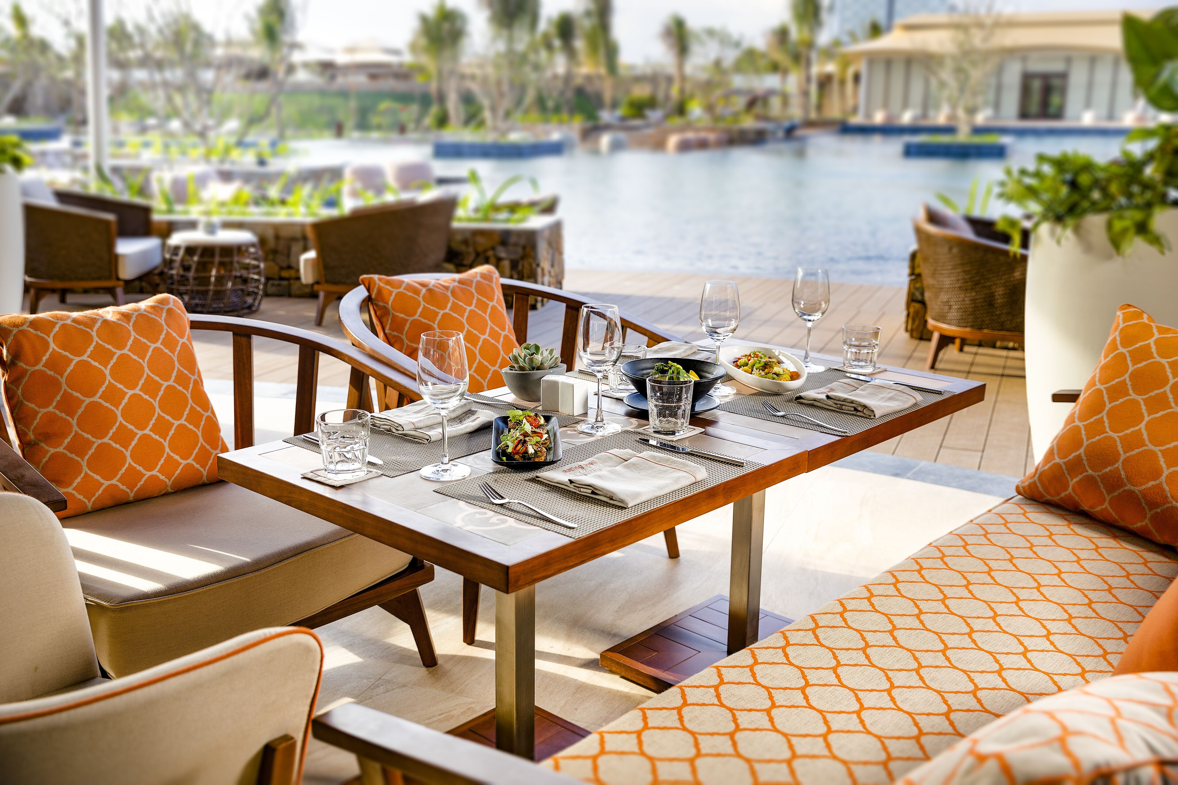 A dining area with a pool view, featuring a table set with plates, glasses, and a bowl of salad.