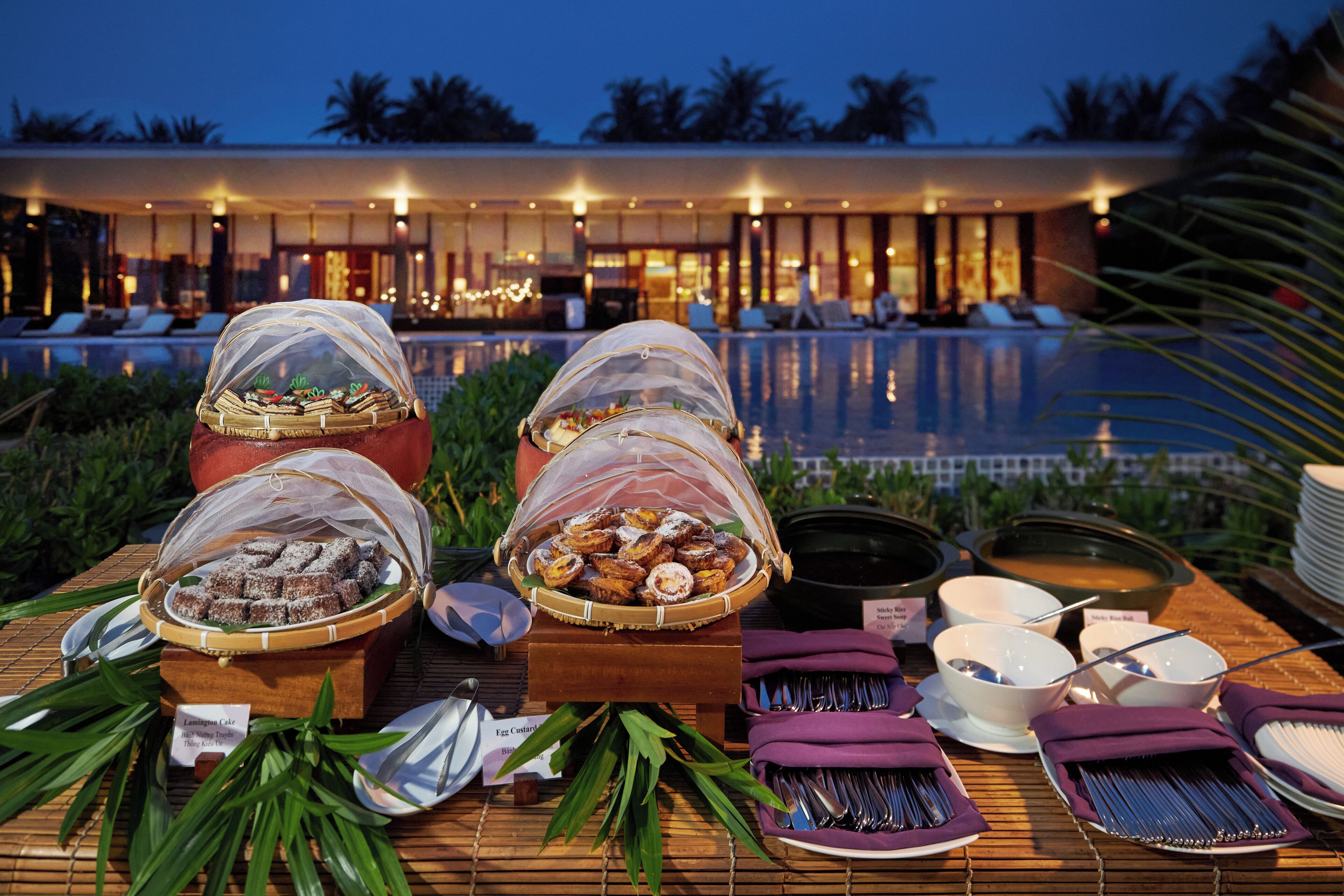 Ein reichhaltiges Buffet mit verschiedenen Speisen und Besteck, im Hintergrund ein Pool und eine tropische Umgebung.