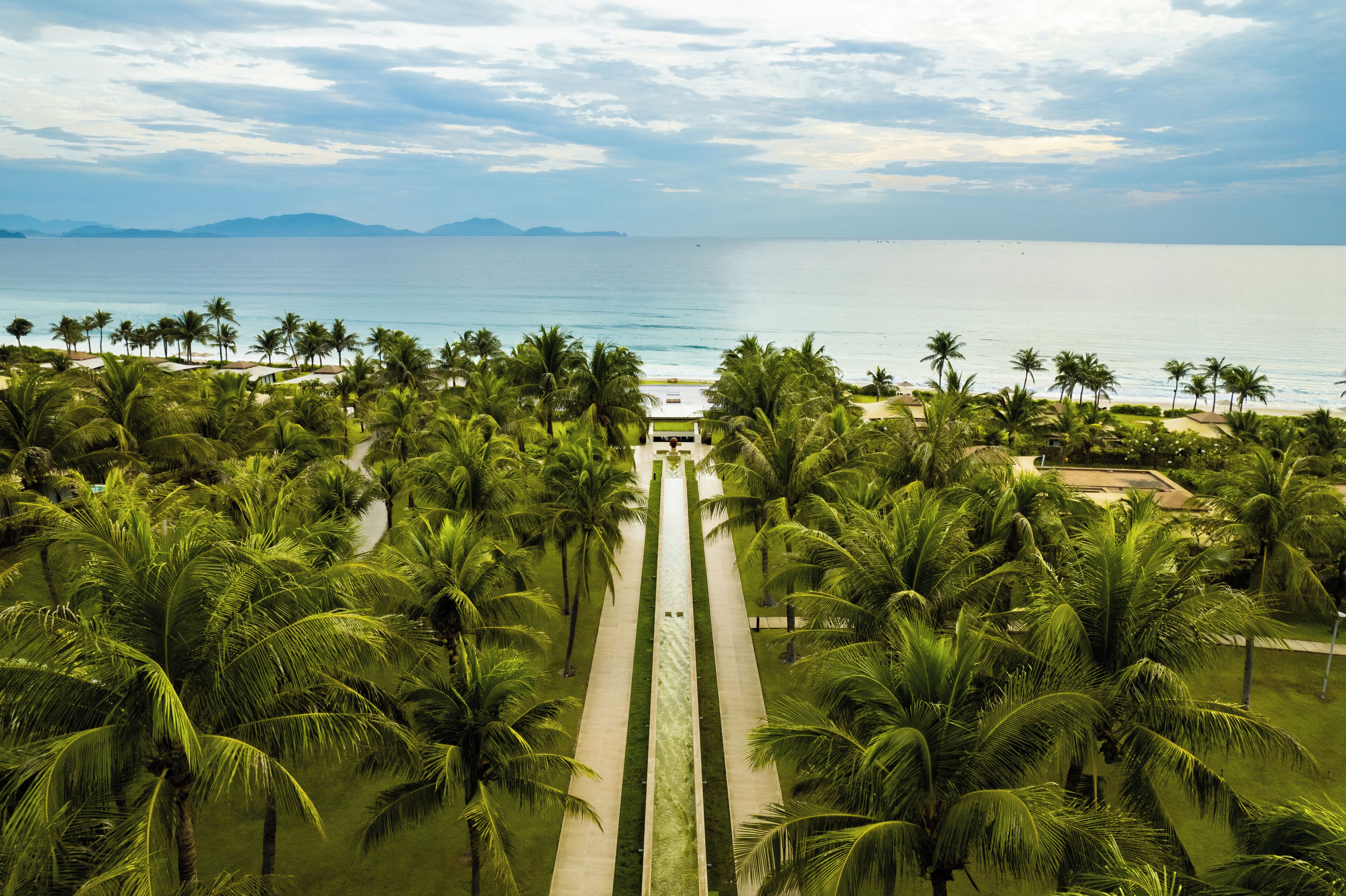 Un resort tropical con un camino bordeado de palmeras que conduce a la playa, con vistas al mar y montañas distantes.