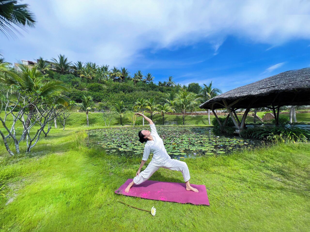 A person performing a yoga pose on a mat in a lush outdoor setting with a pond and palm trees.