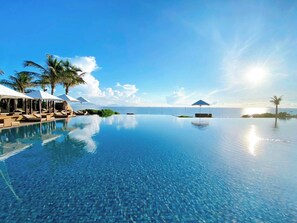 Una piscina infinita con sillones de descanso y sombrillas, palmeras y vista al océano.