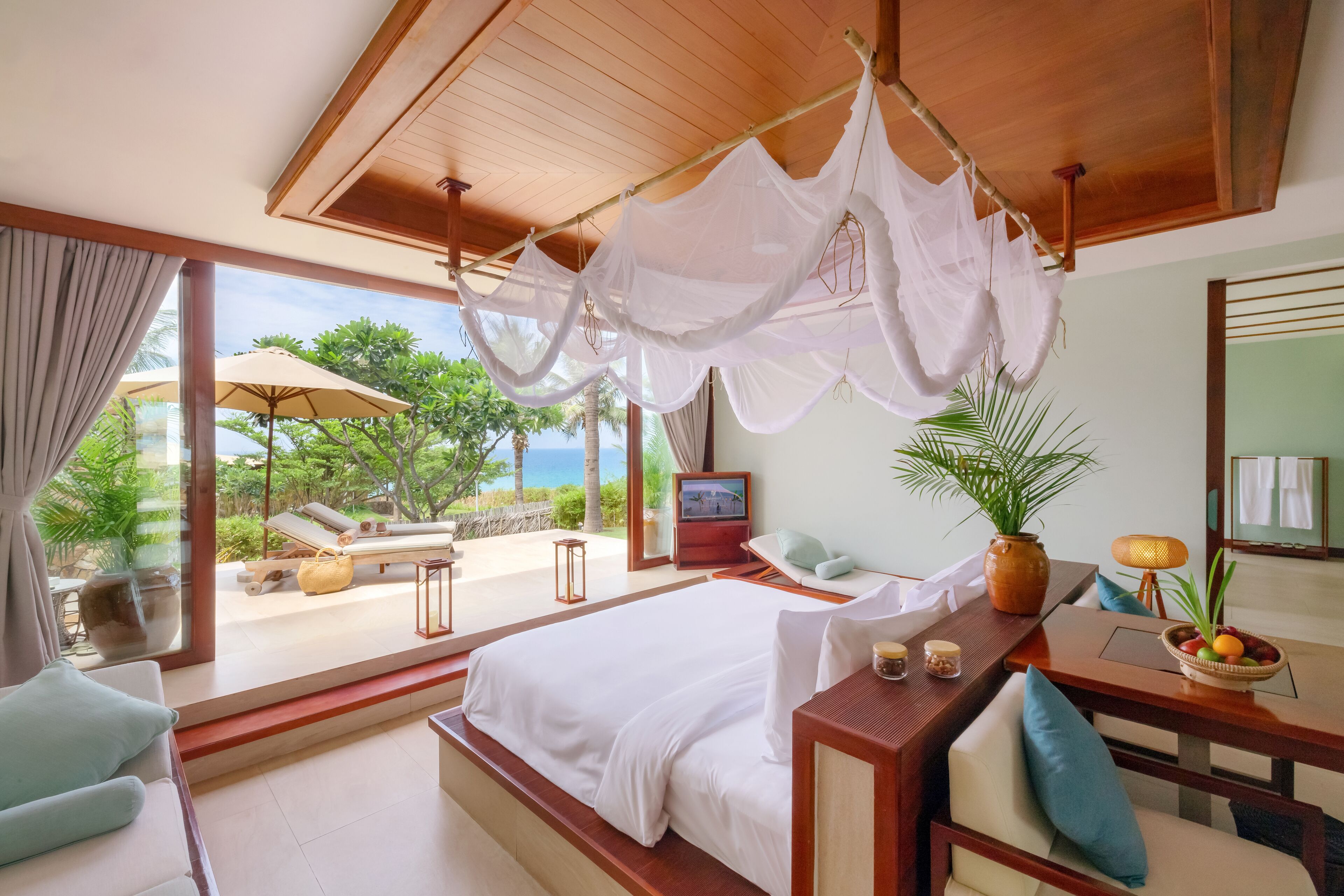 A bedroom with a large bed, a wooden ceiling, a TV, a bedside table, and a view of the outdoors.