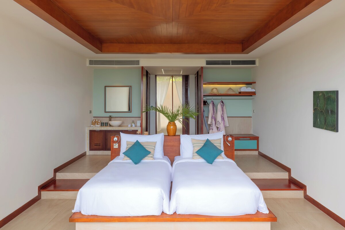 Una cama doble con sábanas blancas y almohadas turquesa, un cabecero de madera, y vistas a un baño con lavamanos y toallero.
