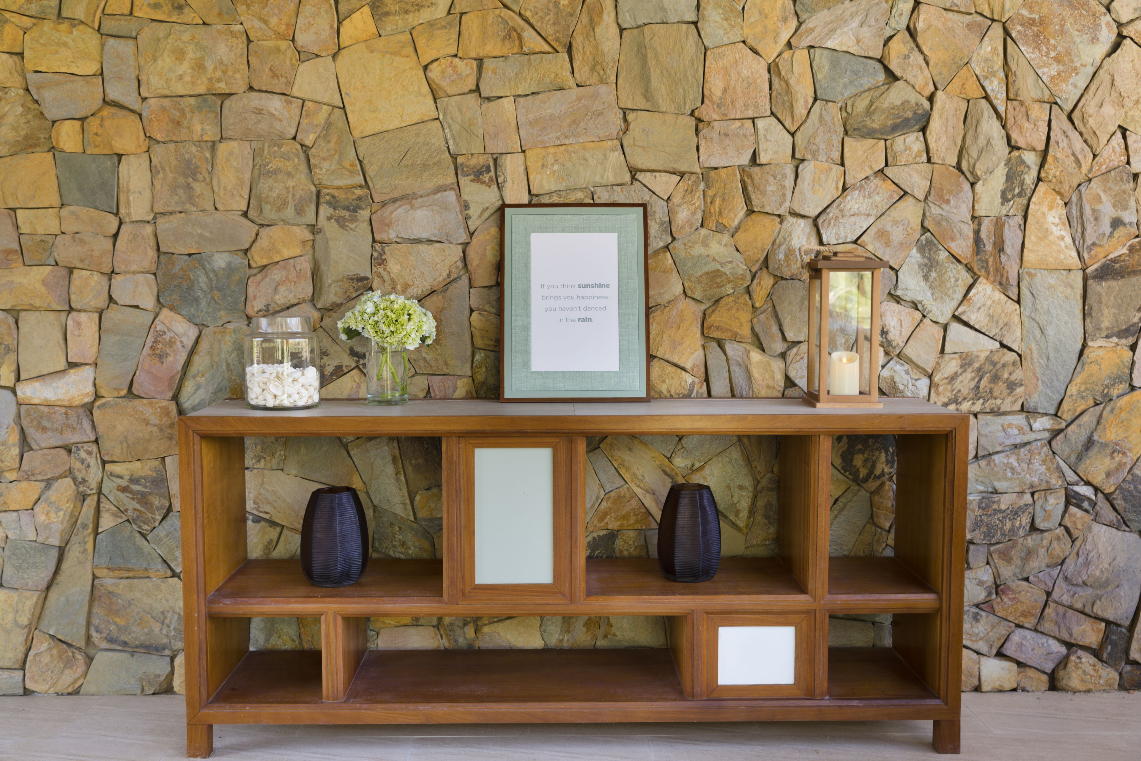 A wooden console table with a stone wall backdrop, featuring a framed inspirational quote, two vases, and a lantern.