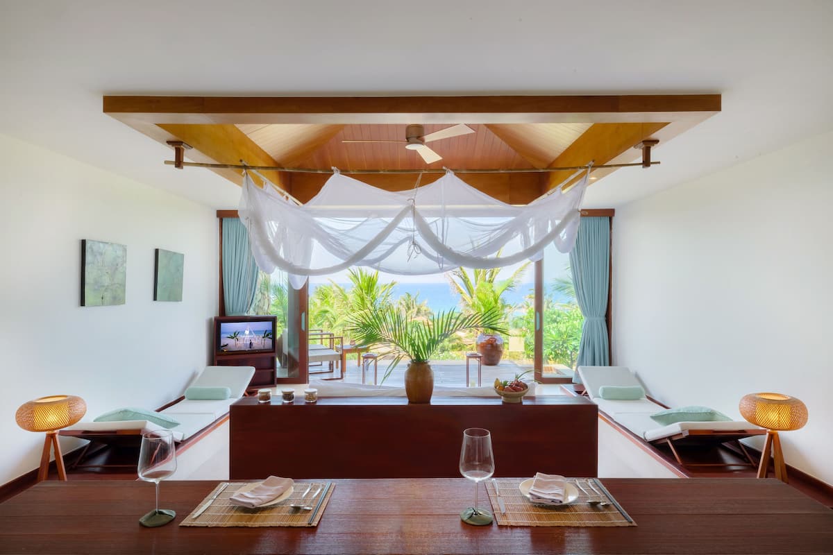 Ein Zimmer mit Holzboden, einem Bett, einem Tisch mit zwei Gläsern, einem Fernseher und Blick auf Palmen und das Meer.