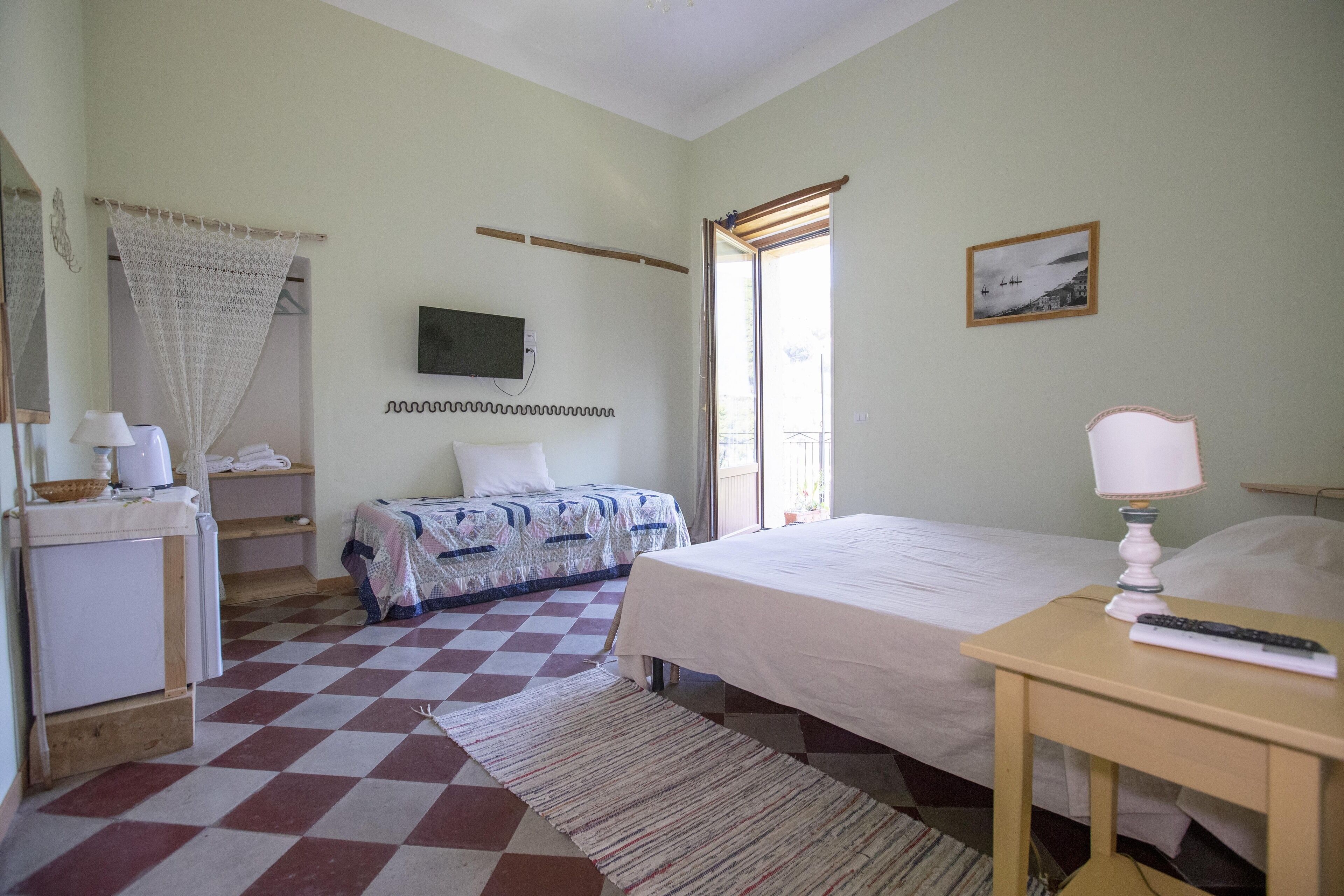 A hotel room with a checkered floor, a bed with a white bedspread, a bedside table with a lamp, a television on the wall, and a window with a view of the sea.