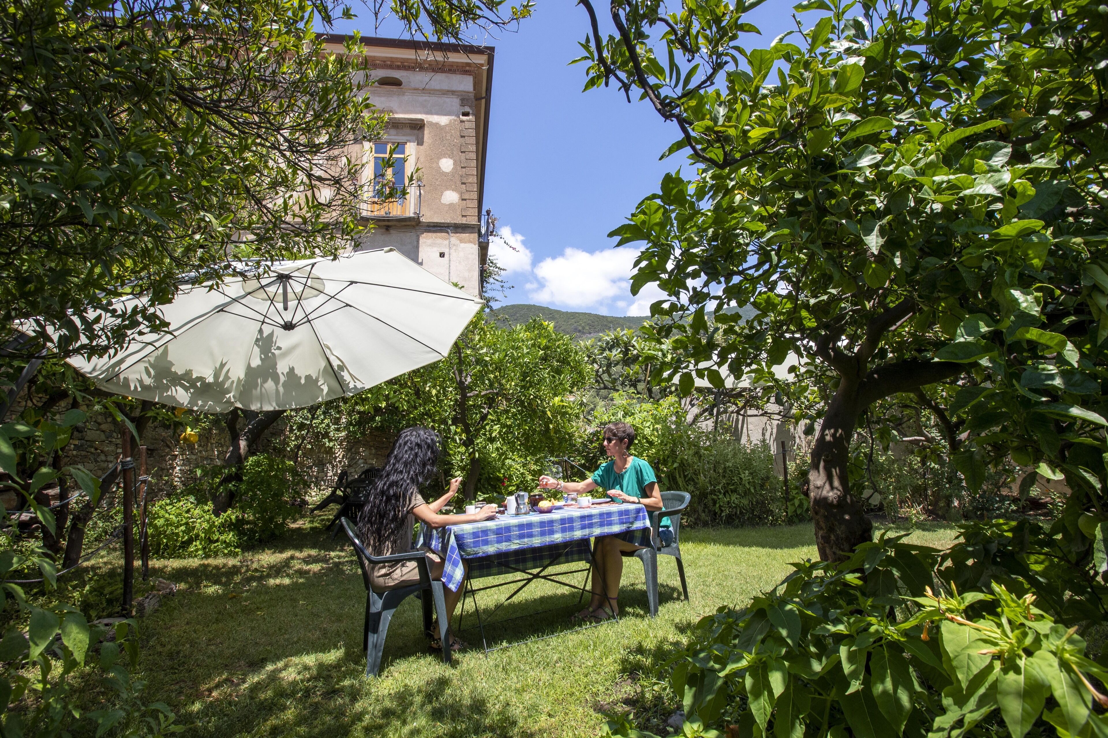Dos personas cenando en una mesa al aire libre con mantel a cuadros, rodeadas de abundante vegetación y un edificio al fondo.