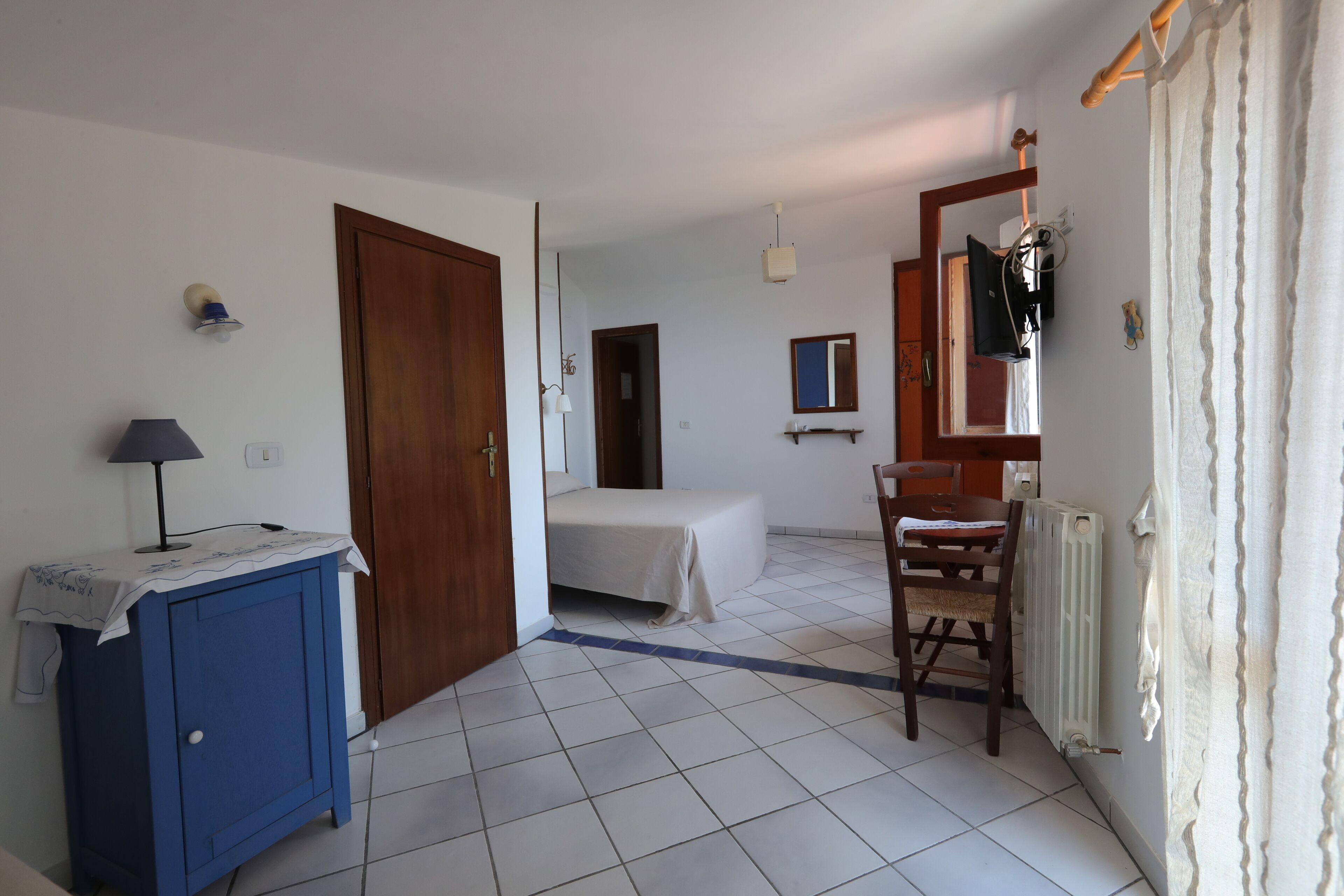 Een kamer met een bed, een bureau met lamp, een stoel, een radiator en een deur naar een andere kamer.