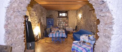 Un comedor rústico con paredes de piedra, techo de madera y suelo de baldosas. Hay mesas con manteles a cuadros azules y blancos, una silla azul y un sofá azul.