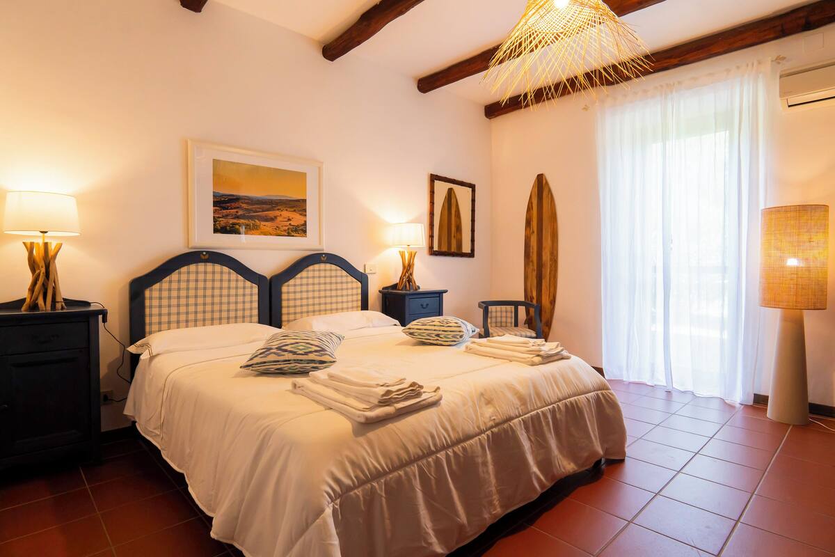 Une chambre à coucher avec un lit, des lampes de chevet, un tableau encadré, une planche de surf et une fenêtre avec des rideaux.