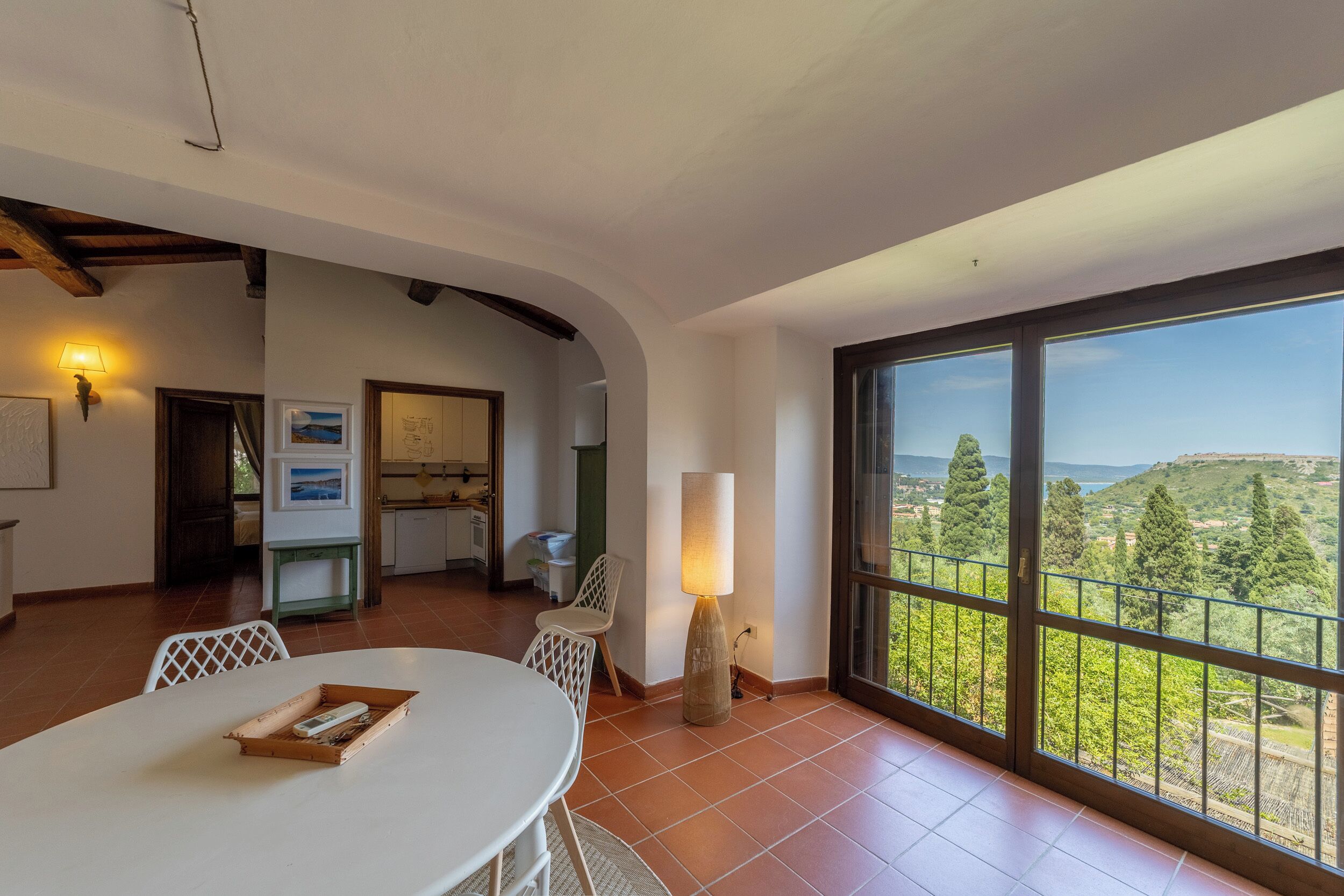 A spacious room with a large window offering a scenic view, a dining area with a table and chairs, and a kitchen in the background.