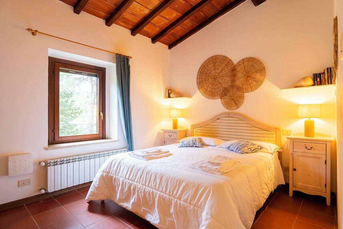 Une chambre à coucher comprenant un lit, deux tables de chevet, une fenêtre avec des rideaux, un radiateur et un élément de décoration murale.