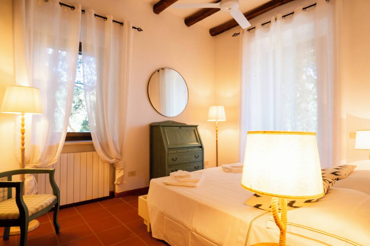 Une chambre à coucher avec un lit, deux lampes de chevet, une commode, une chaise et une fenêtre avec des rideaux.