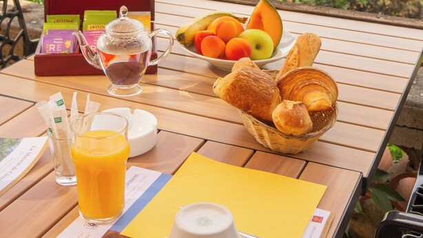 A breakfast table with a basket of croissants, a fruit bowl, a teapot, and a juice glass on a balcony overlooking a scenic countryside. - Hotel Le Relais Des Deux Vallees (Salvagnac)