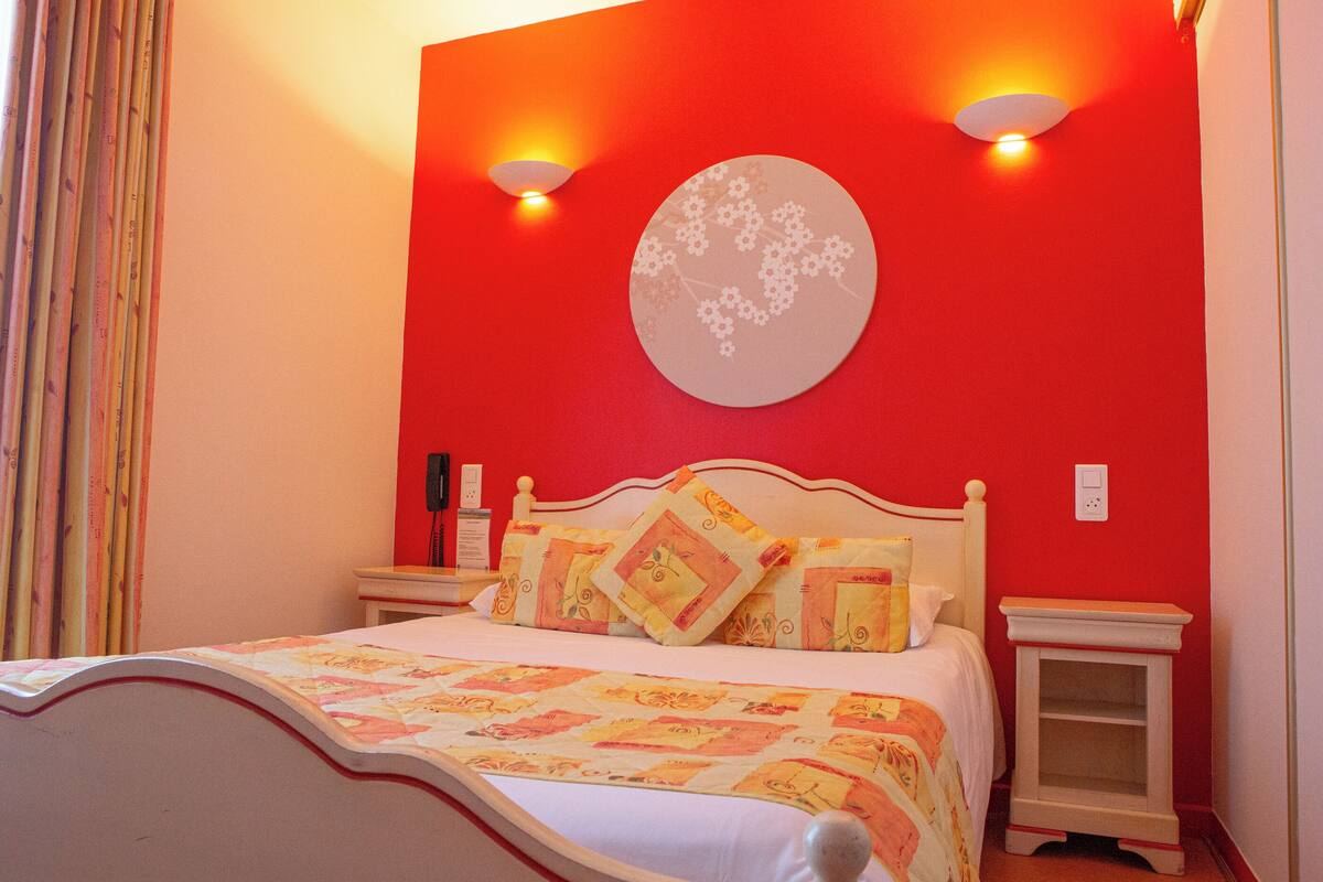 A bedroom with a red wall, a bed with a floral bedspread, two bedside tables, and a wall-mounted light fixture.