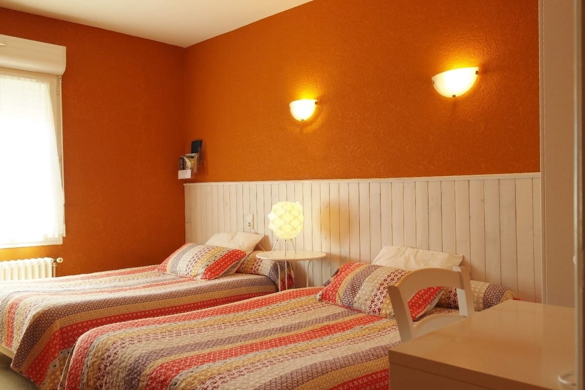 Une chambre avec deux lits, une tête de lit blanche et deux lampes fixées au mur.