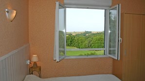 Et soveværelse med seng, et natbord med lampe, et vindue med åben udsigt over et grønt landskab og en træskab.
