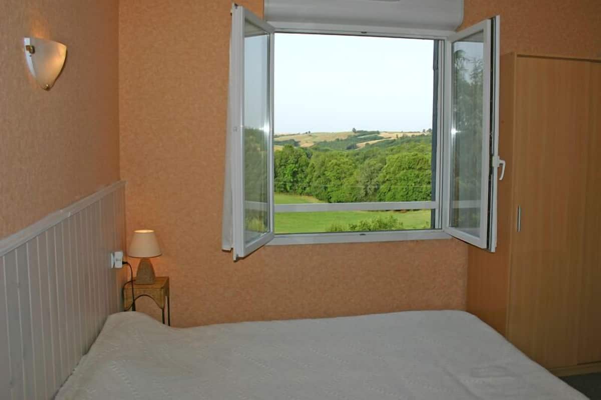 Une chambre à coucher avec un lit, une table de chevet avec une lampe, une fenêtre donnant sur un paysage verdoyant, et une armoire en bois.