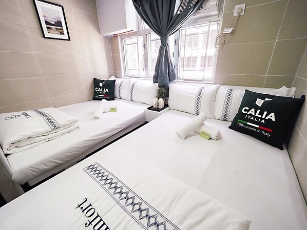 A hotel room with two beds, each with white linens and pillows featuring the brand "Callia Italia." There is a window with curtains, a framed picture on the wall, and a small table with toiletries. - Joyous Guest House (Kowloon)
