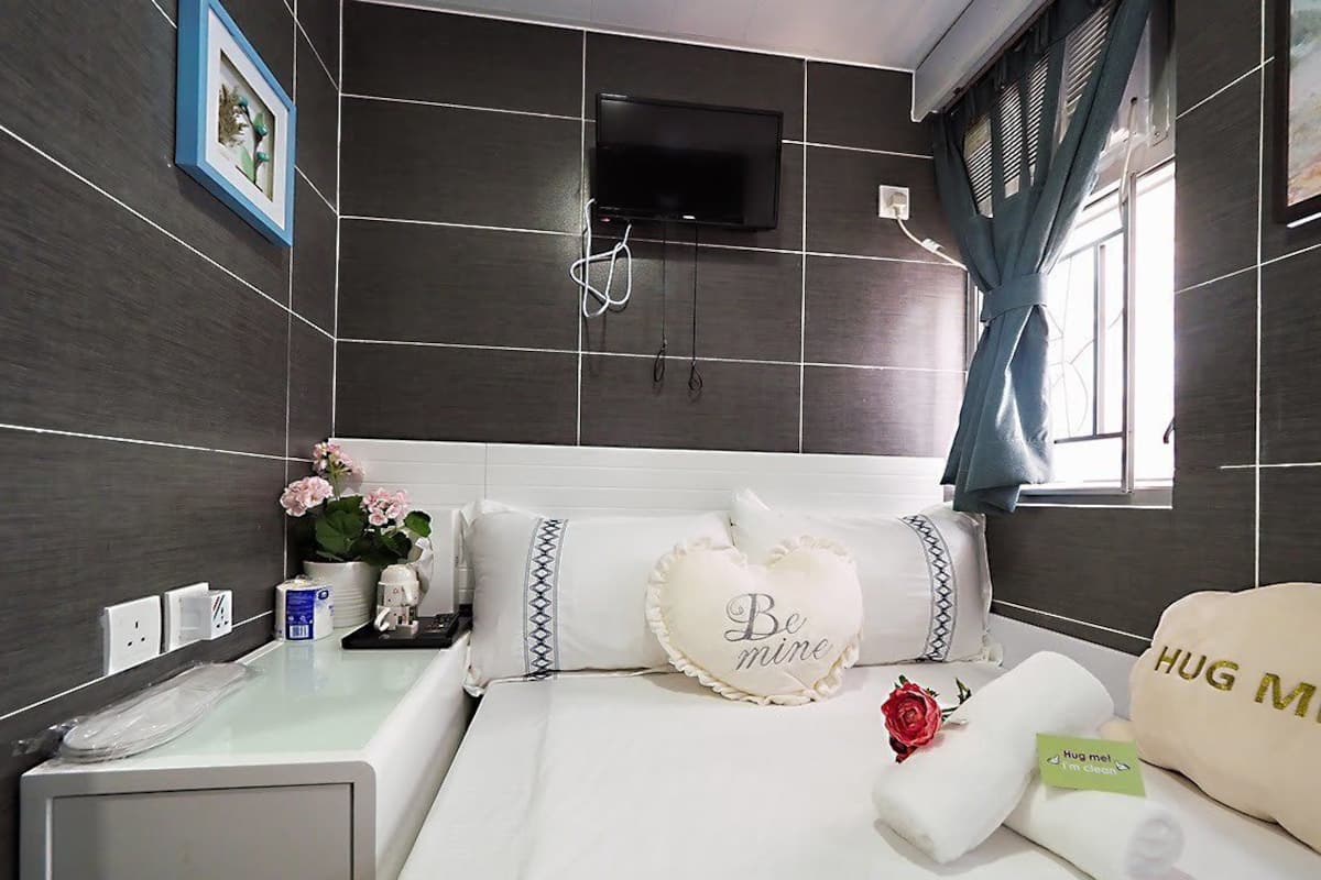 A hotel room with a large bed, a flat-screen TV mounted on the wall, a bedside table with a lamp, and a window with curtains.