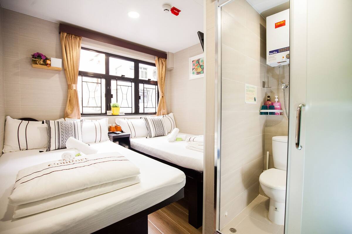 A hotel room with two beds, a window with curtains, a shower area, and a toilet.
