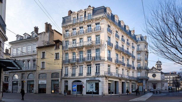 A multi-story hotel with balconies, a ground-level shop, and a church in the background. - Hôtel de l'Europe Grenoble Hyper Centre (Grenoble)