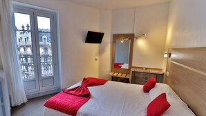 A hotel room with a bed, a TV, a mirror, and a window with a view of buildings. - Hôtel de l'Europe Grenoble Hyper Centre (Grenoble)