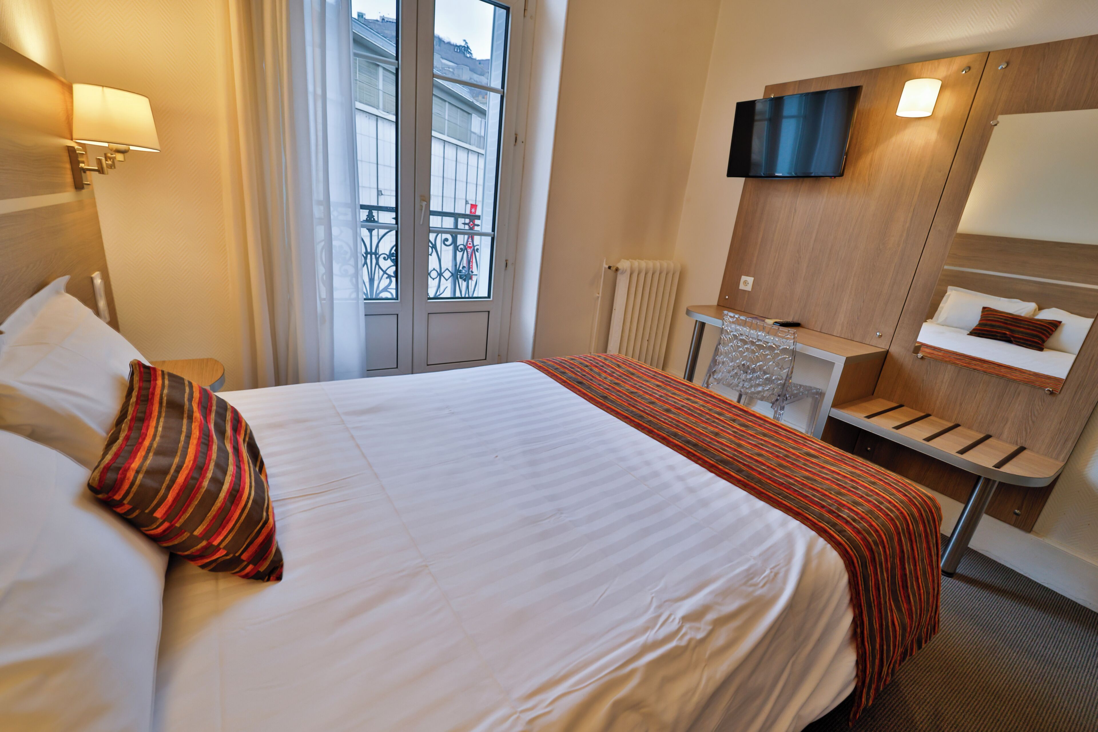 A hotel room with a large bed, a TV mounted on the wall, a small desk, and a window with a view of buildings.