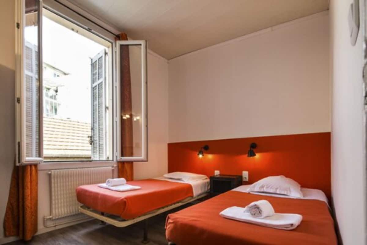 A hotel room with two beds, a window with blinds, a radiator, and a wall-mounted light fixture.