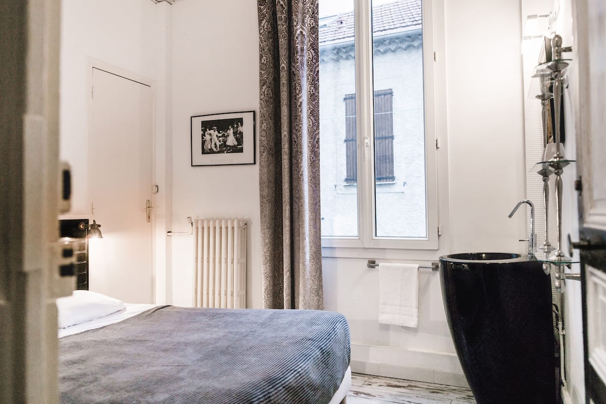 A bedroom with a bed, a window with curtains, a radiator, and a bathroom with a sink and mirror.