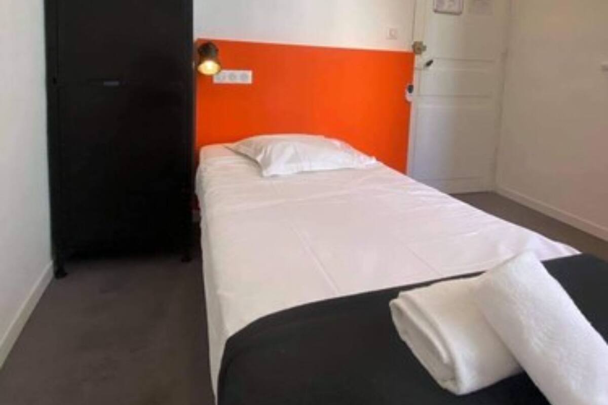 A single bed with white bedding, an orange headboard, and a black side table with a folded white towel.