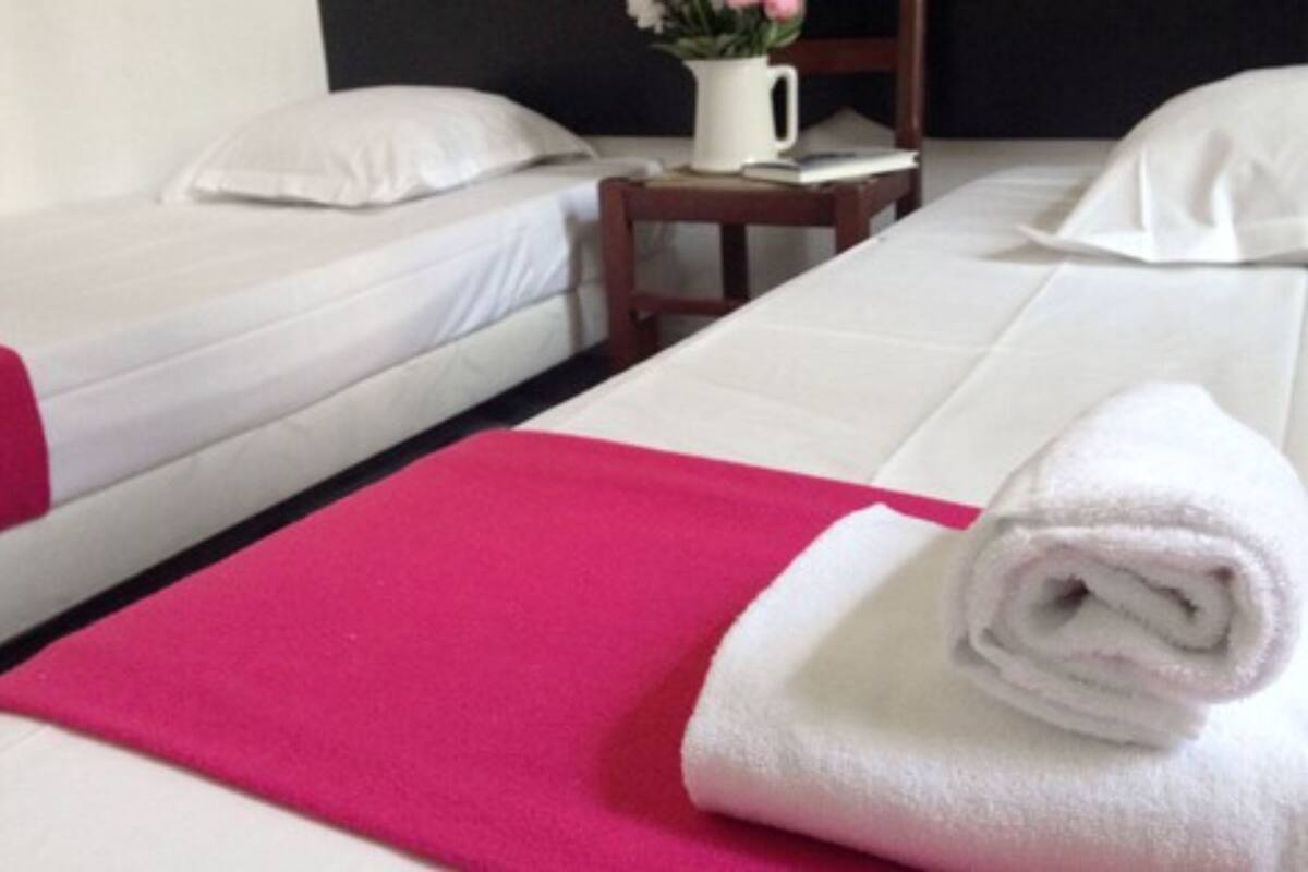A hotel room with two beds, each with white linens and pillows. There is a pink towel on the bed in the foreground and a white towel on the bed in the background. A small wooden nightstand with a white pitcher and a book is visible between the beds.