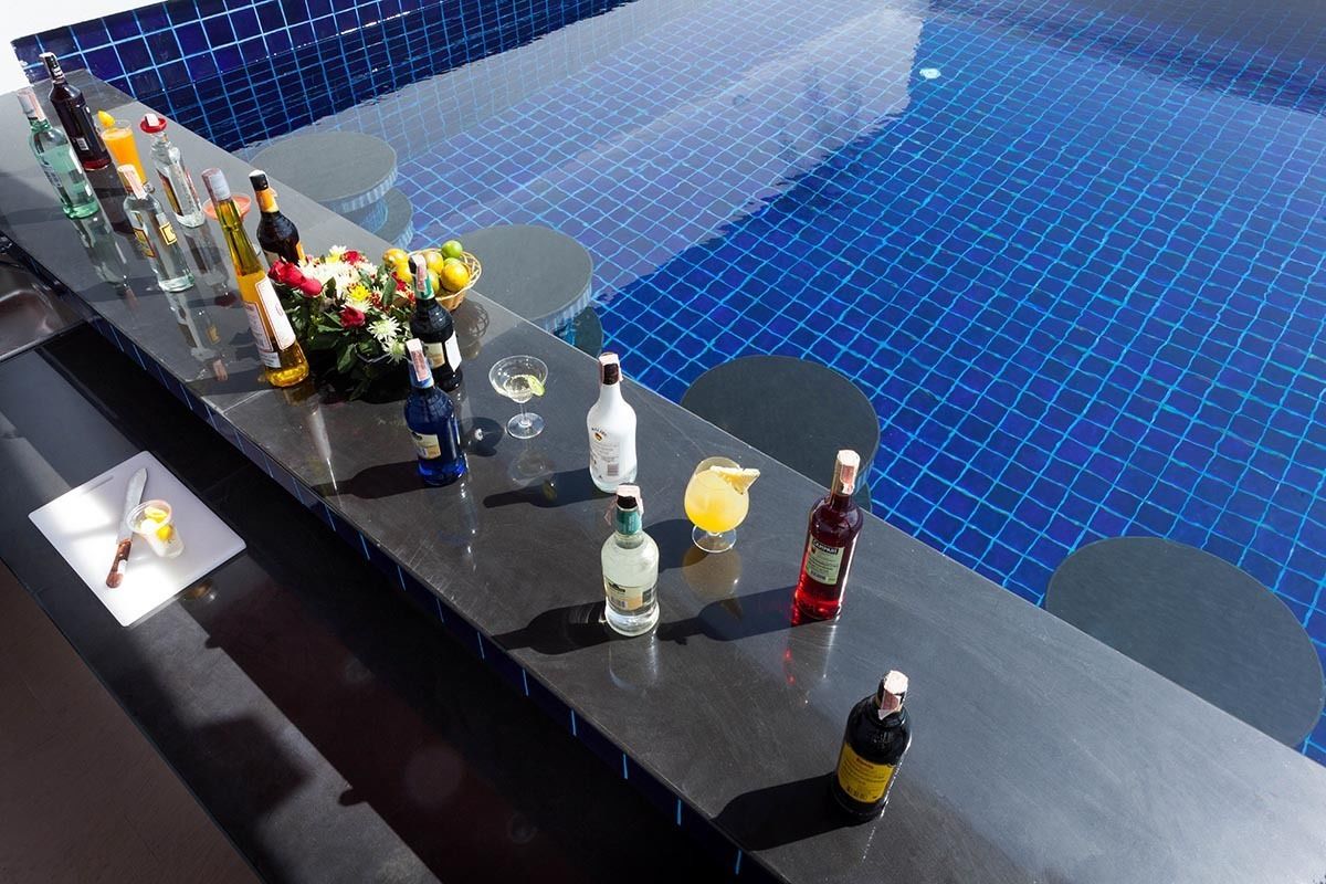 A poolside bar with various bottles and glasses, a knife on a cutting board, and a bowl of fruit.