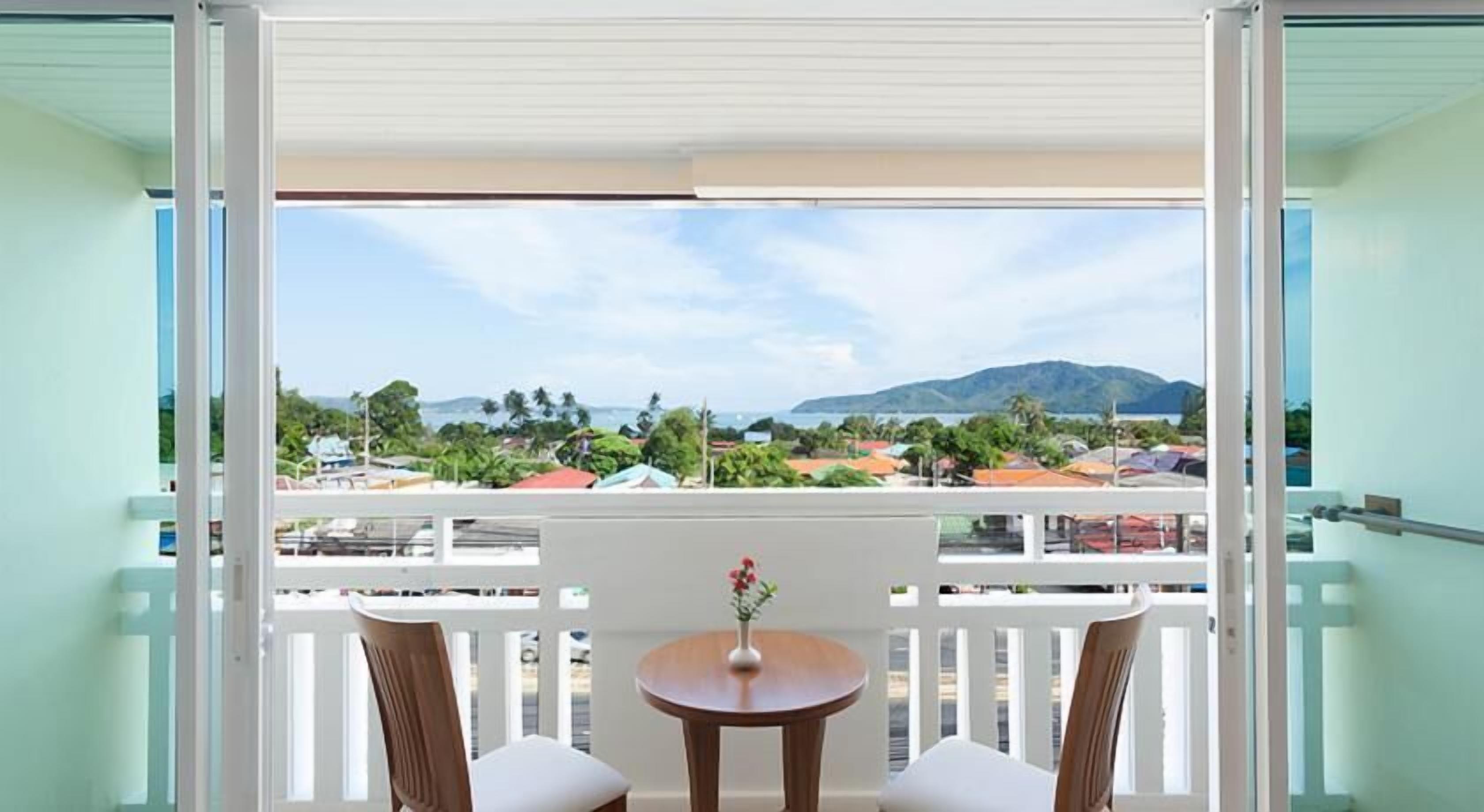 A balcony with a table and chairs, a vase with flowers, and a view of a coastal area with buildings and mountains.