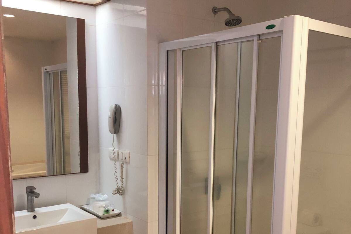 Un baño con lavamanos blanco, espejo, teléfono y ducha con mampara de vidrio.