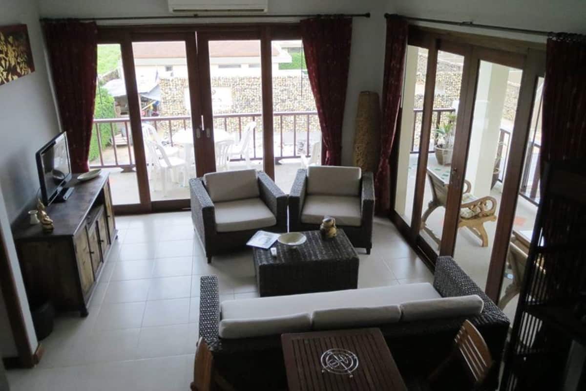 A spacious living area with a sofa, armchairs, and a coffee table. There is a television on a cabinet, and the room has large sliding glass doors leading to an outdoor patio.