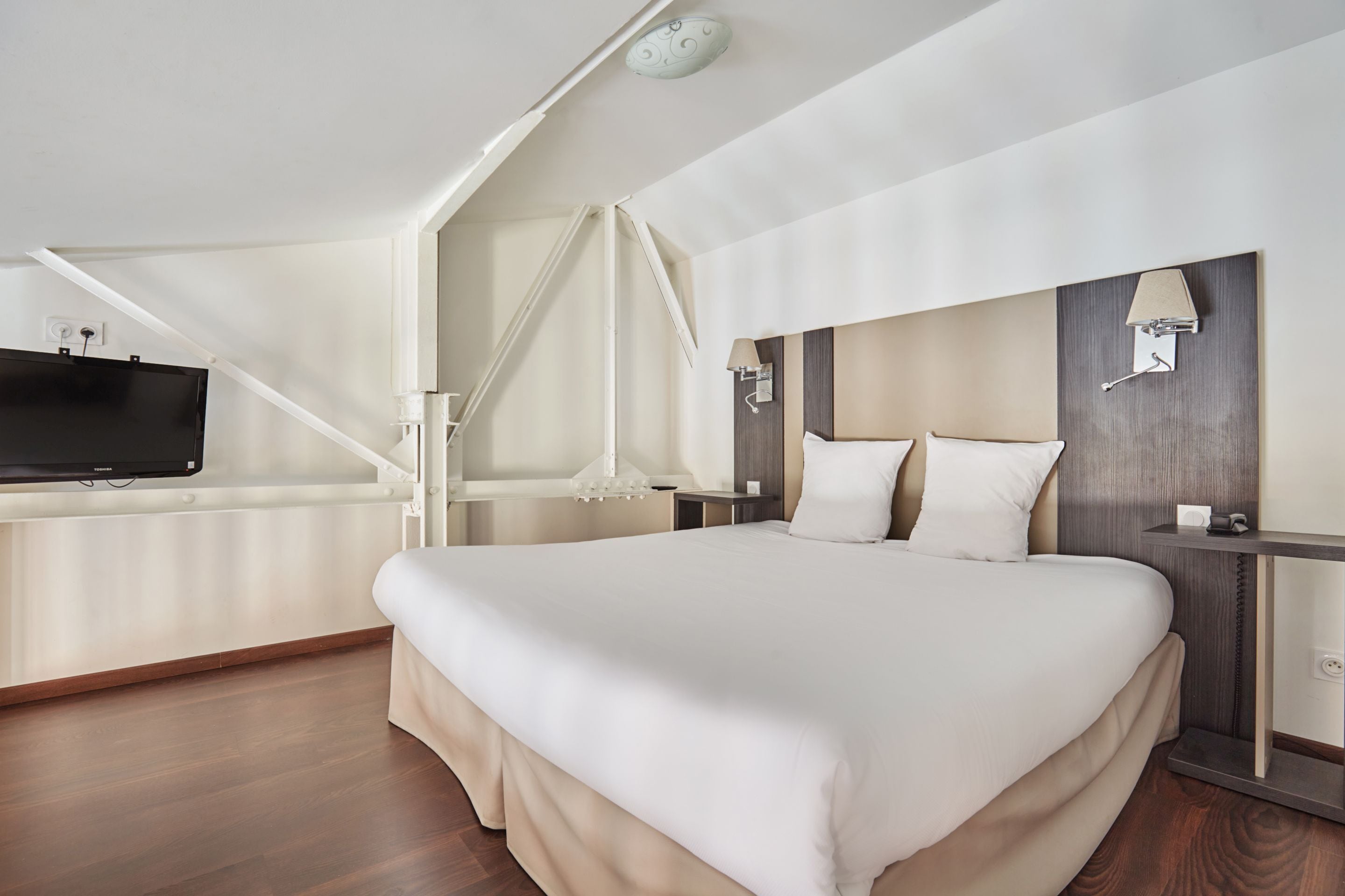 A modern hotel room with a large bed, a TV mounted on the wall, a desk with a lamp, and a wooden floor.